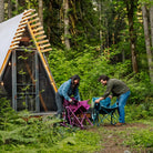 Two people fold up a berry and a teal Kickback Insulated Rocker outside an A-frame cabin surrounded by forest.