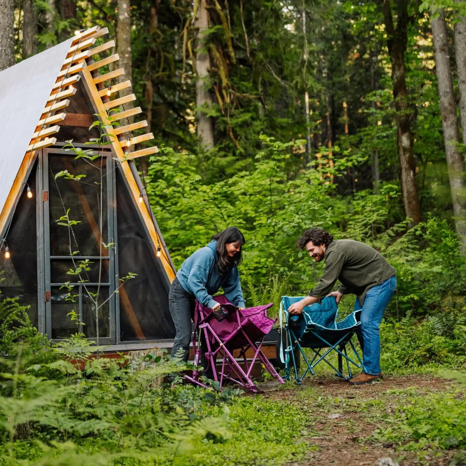 Two people fold up a berry and a teal Kickback Insulated Rocker outside an A-frame cabin surrounded by forest.