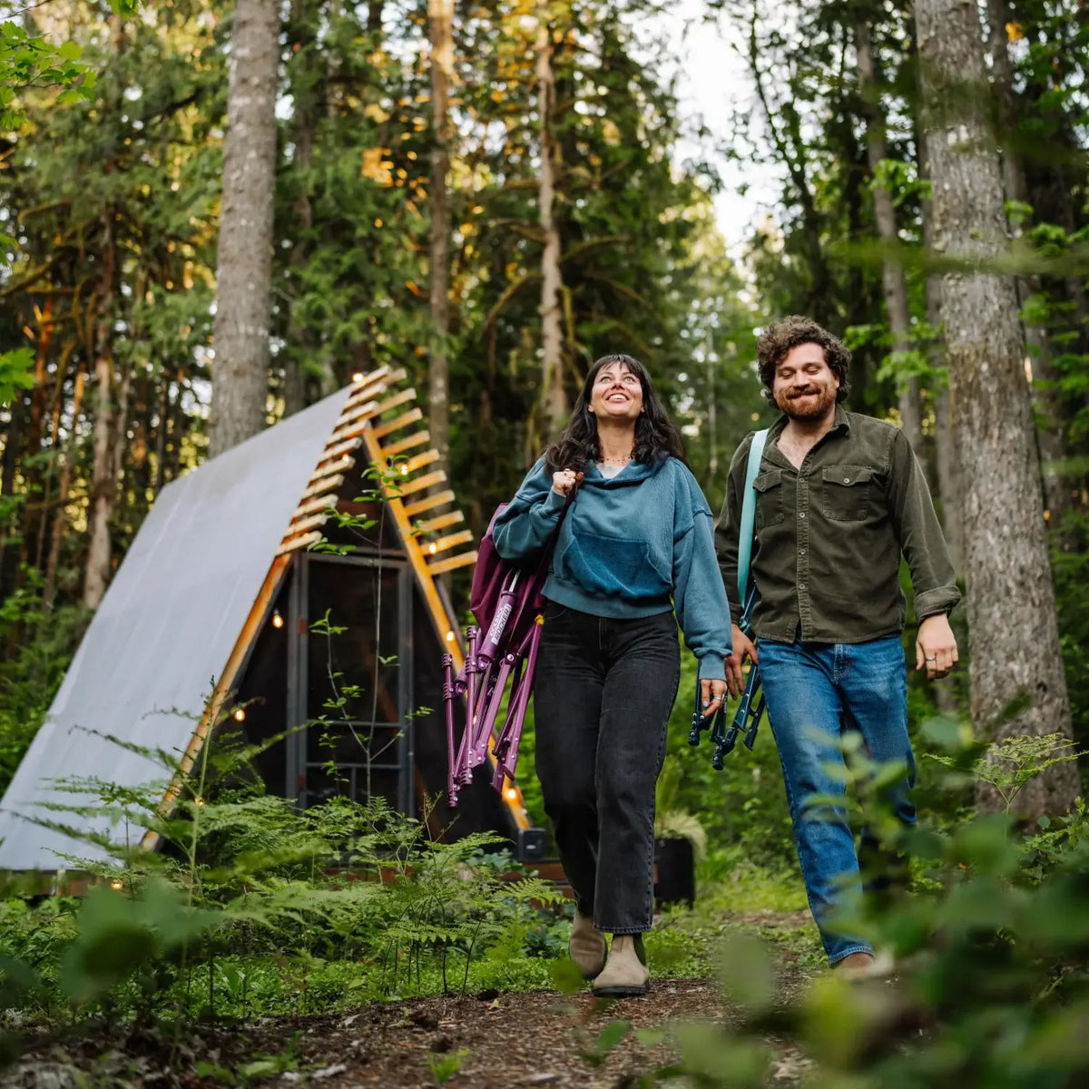 Man and woman walk through the woods carrying a folded berry and a folded teal Kickback Insulated Rocker, with an A-frame cabin behind them.