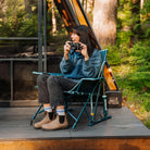 Woman photographs something off-camera while seated in a teal Kickback Insulated Rocker on a deck outside an A-frame cabin.