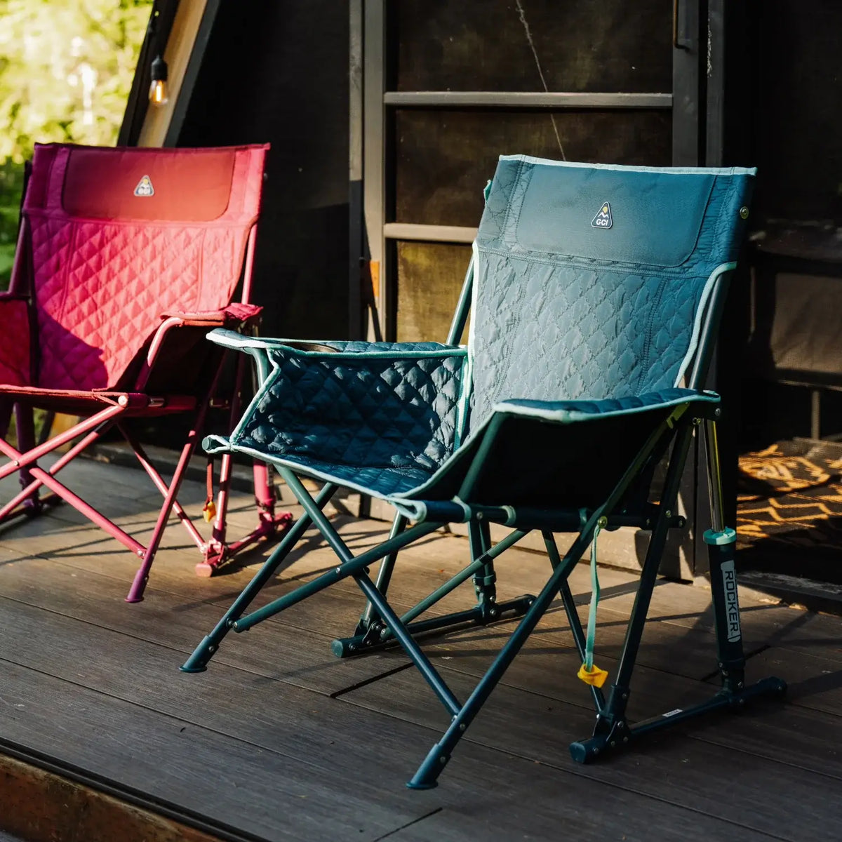 A teal Kickback Insulated Rocker with quilted seating sits on a wooden deck beside a berry chair, near an A-frame cabin in the woods.