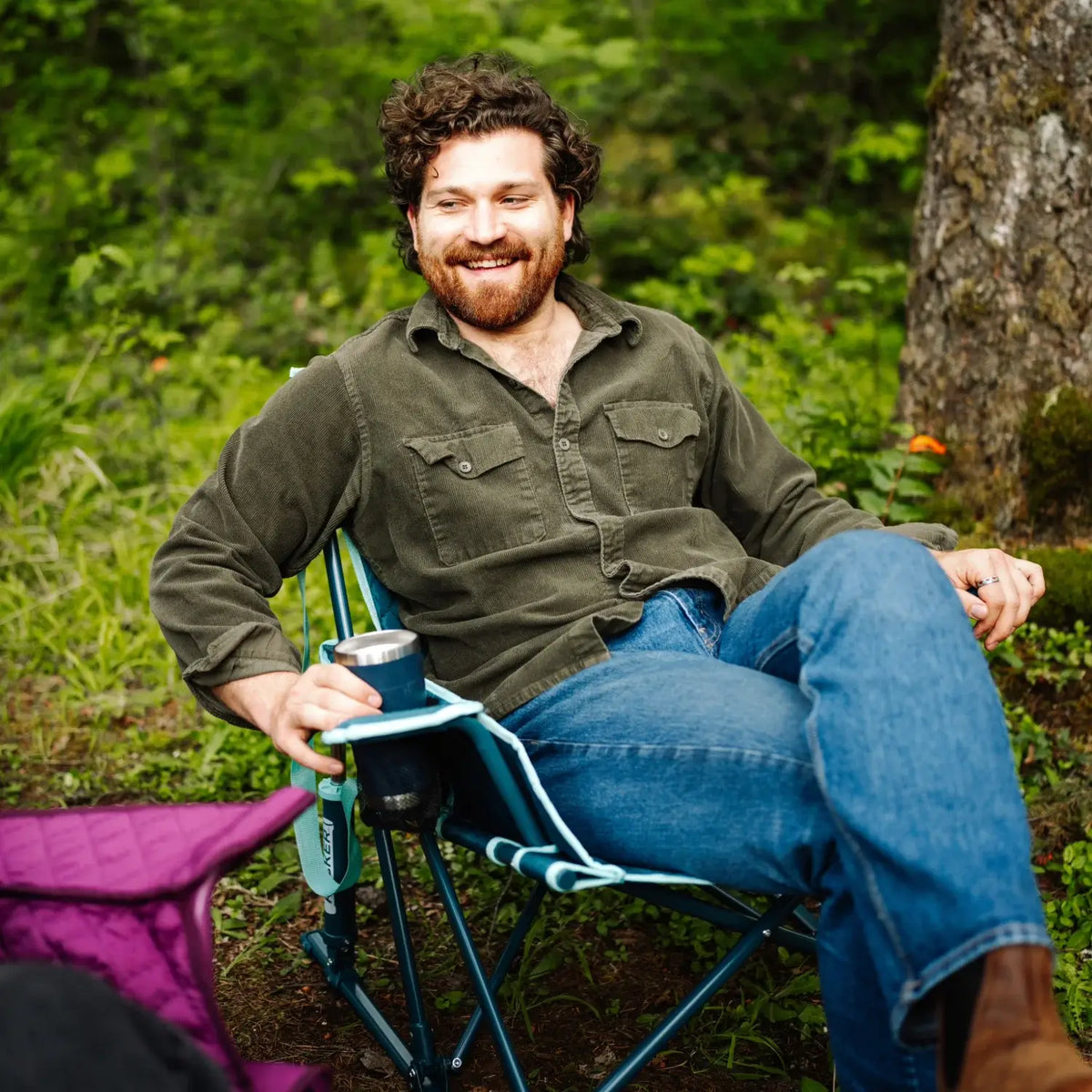 Man relaxes in a teal Kickback Insulated Rocker in the woods, resting a metal cup in the built-in cup holder, with a berry chair beside him.
