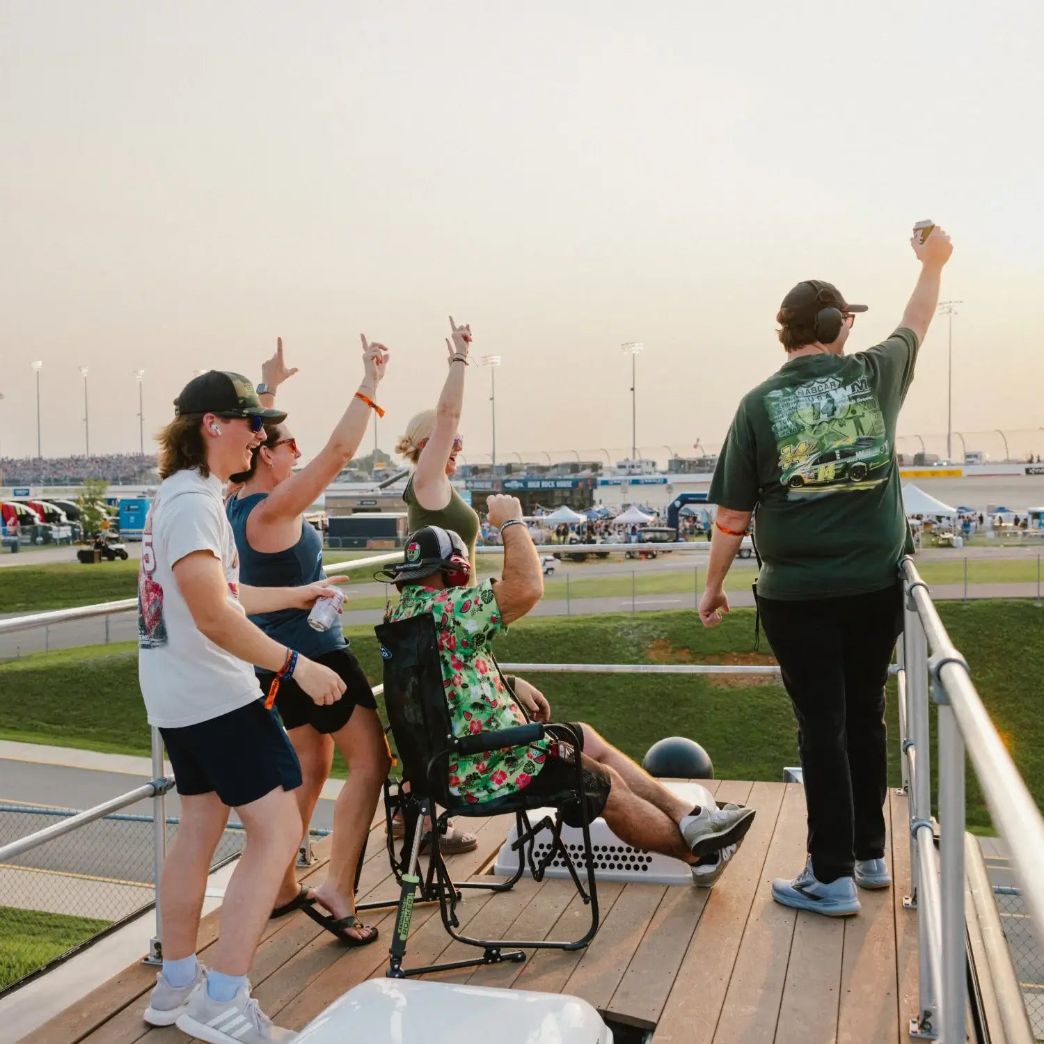 Group of NASCAR fans cheering from a trackside deck at sunset during a race.