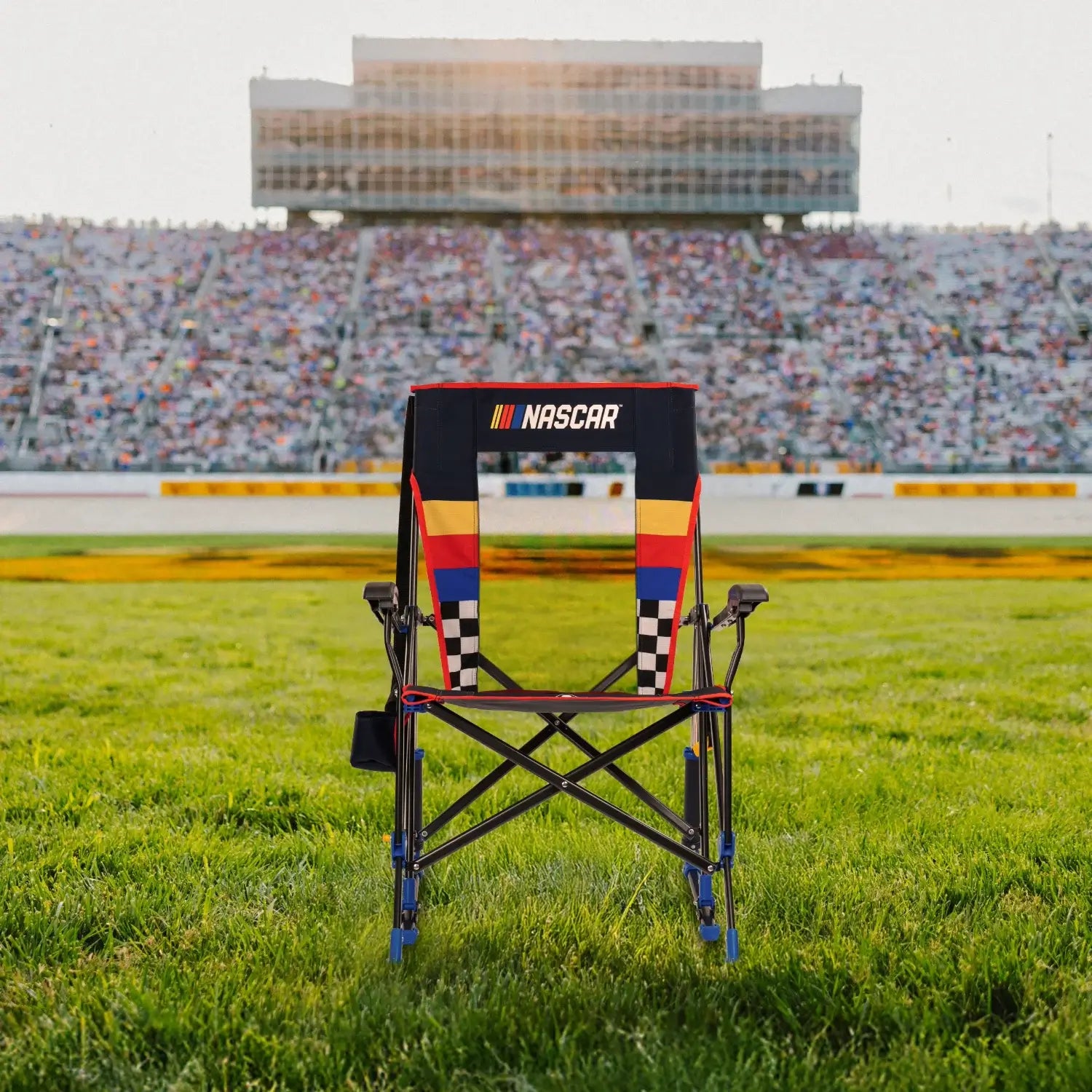 A Roadtrip Rocker NASCAR chair sitting on a patch of grass in front of a racecar track. 