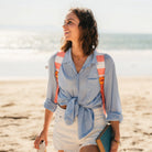 Smiling woman walks along the beach with a coral-striped GCI SideKick beach chair strapped to her back like a backpack.