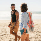 Woman carries a folded coral-striped GCI SideKick beach chair on her back while walking alongside a friend on the beach.
