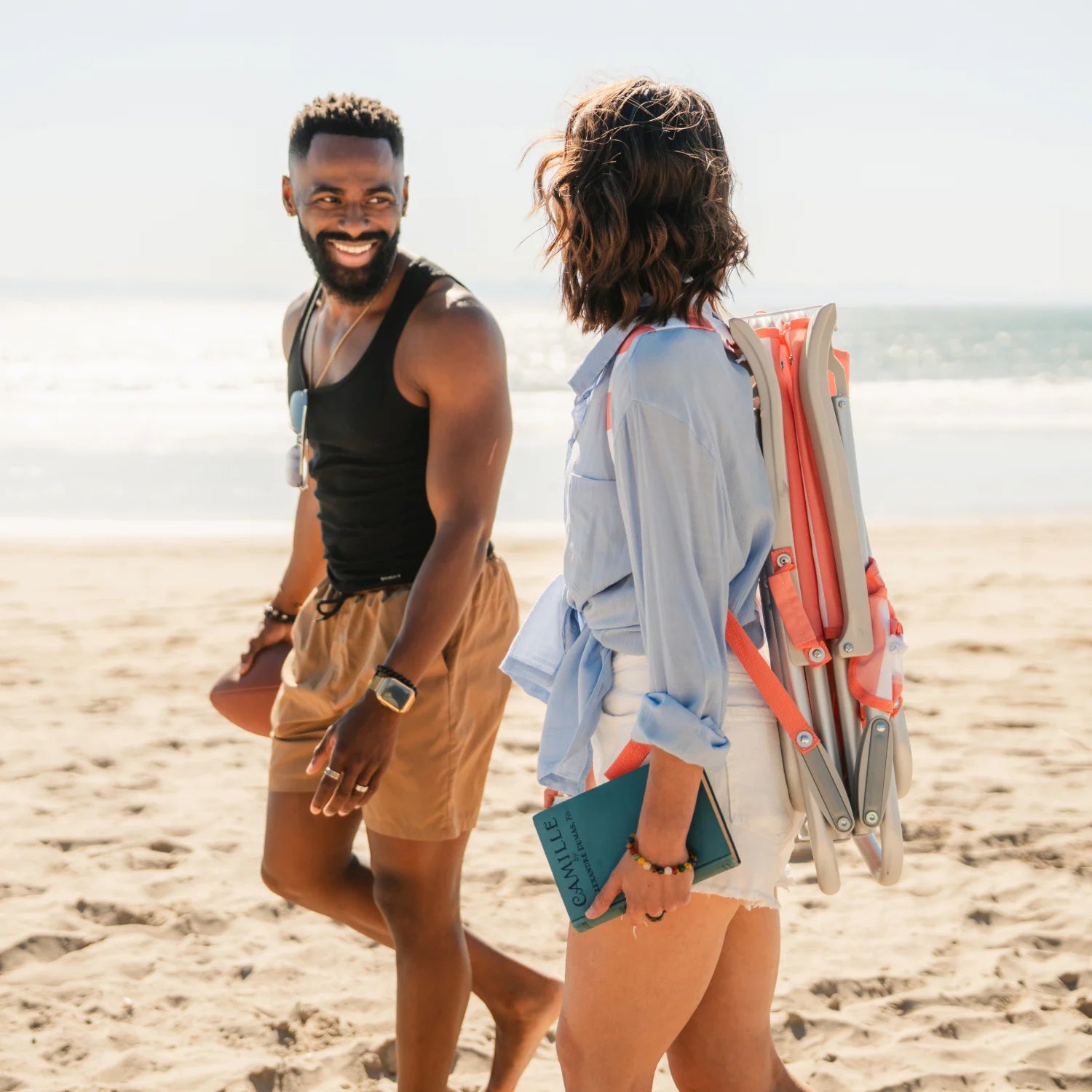 Woman carries a folded coral-striped GCI SideKick beach chair on her back while walking alongside a friend on the beach.