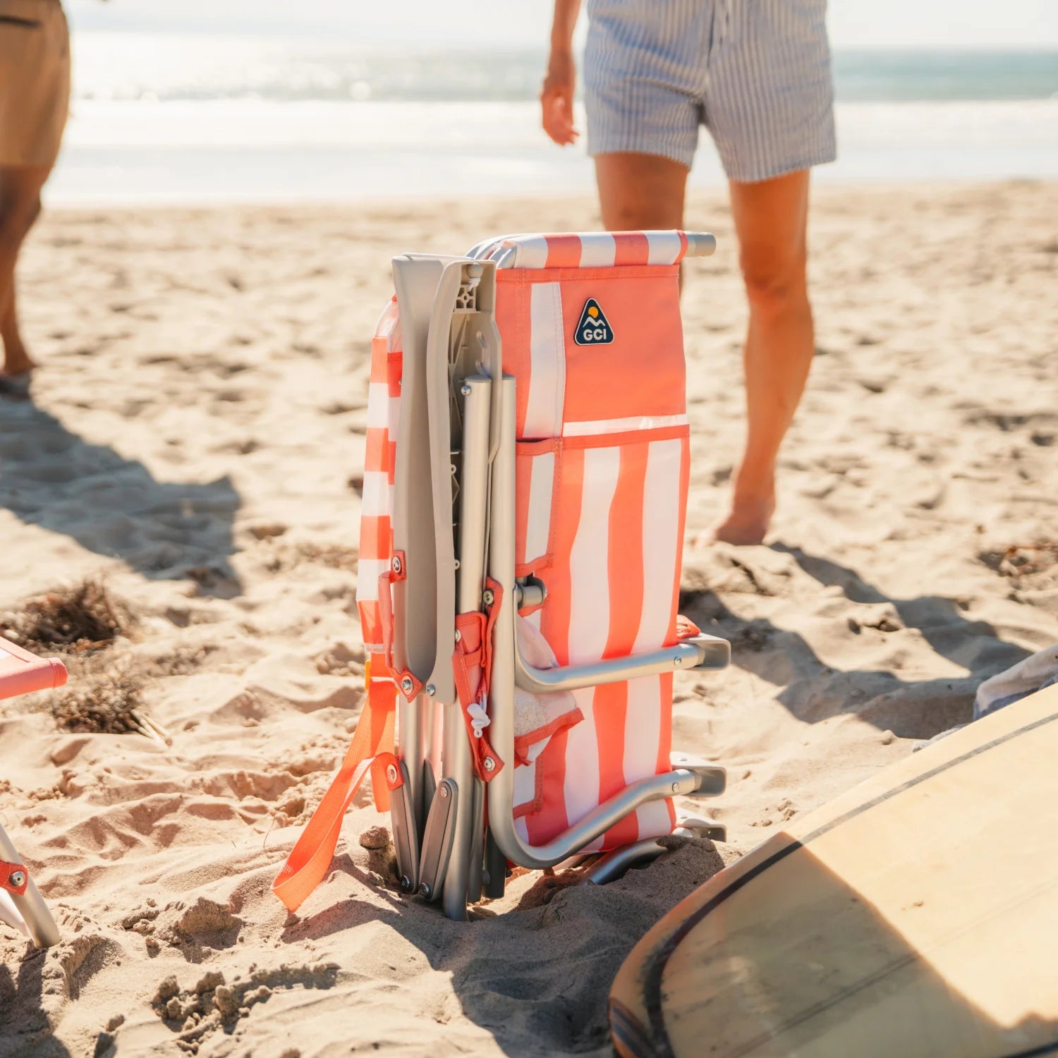 Folded coral-striped GCI SideKick beach chair stands upright in the sand next to a surfboard at the shoreline.
