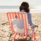 Woman relaxes in a coral-striped GCI SideKick beach chair facing the ocean showing the mesh back panel and logo.