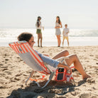 Smiling woman reclines in a coral-striped GCI SideKick beach chair with friends chatting near the water in the background.