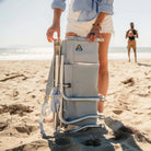 Folded heathered light blue GCI SideKick beach chair stands upright in the sand held by a woman.