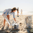 Woman setting up the SideKick heathered light blue beach chair with armrest storage pockets on the sand.