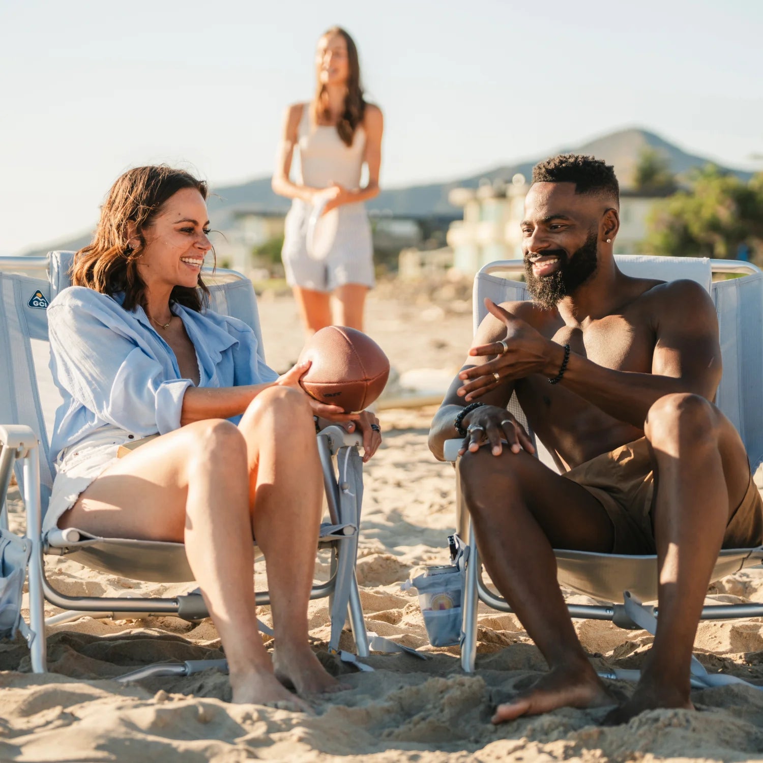 Two people chat while seated in heathered light blue GCI SideKick beach chairs on the sand.