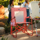GCI Outdoor Stowaway Rocker in Coral Mosaic, front right angle view, at an outdoor gathering with a charcuterie board nearby.