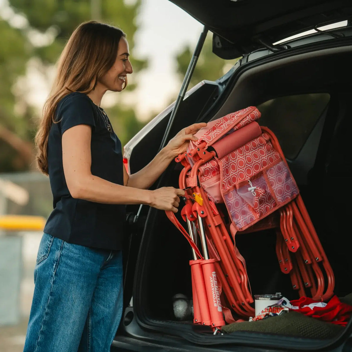 Woman loading a folded GCI Outdoor Stowaway Rocker in Coral Mosaic into a car trunk.