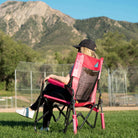 Back view of a woman seated in a pink flame waves Stowaway Rocker on a grass field with mountains in the background.