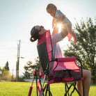 Woman seated in a pink flame waves Stowaway Rocker lifts a baby into the air on a sunny day in a grassy field.