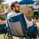 A man sits in a blue GCI outdoor chair at a sports field, holding a drink and watching a game. The chair has a breathable mesh back and side storage pockets.