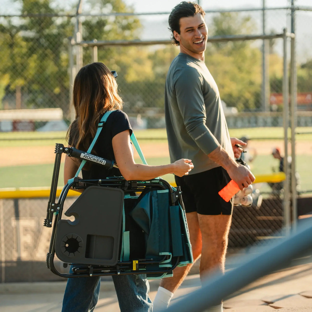 A man and woman walking next to a baseball field while a woman carries a teal Stowaway Rocker with Side Table with a shoulder strap. 