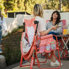 Two women sitting in a coral Stowaway Rocker with one of the chairs having a clipped coral Sunshade Accessory attached. 