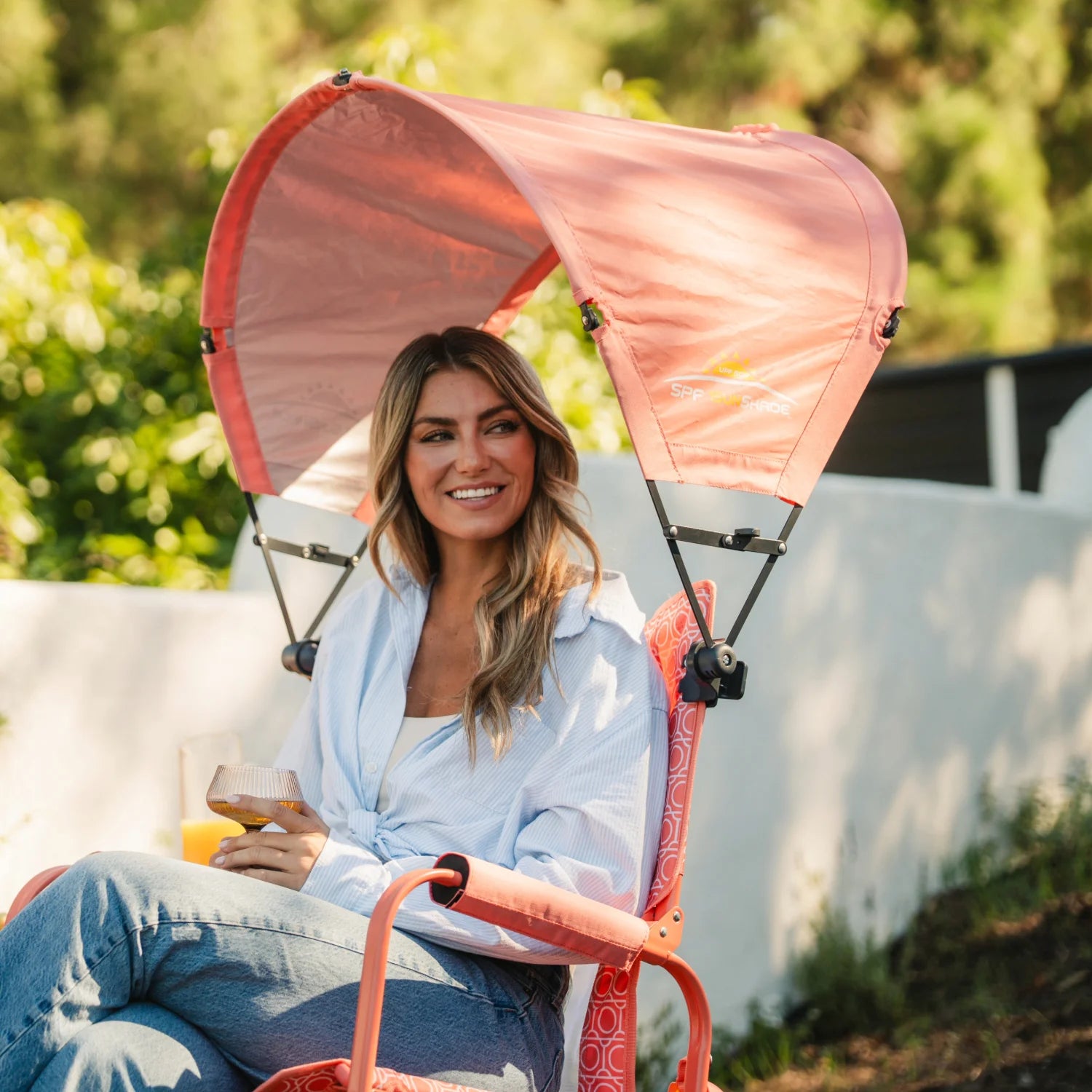 A woman sitting in a coral Stowaway Rocker with a coral Sunshade Accessory attached. 