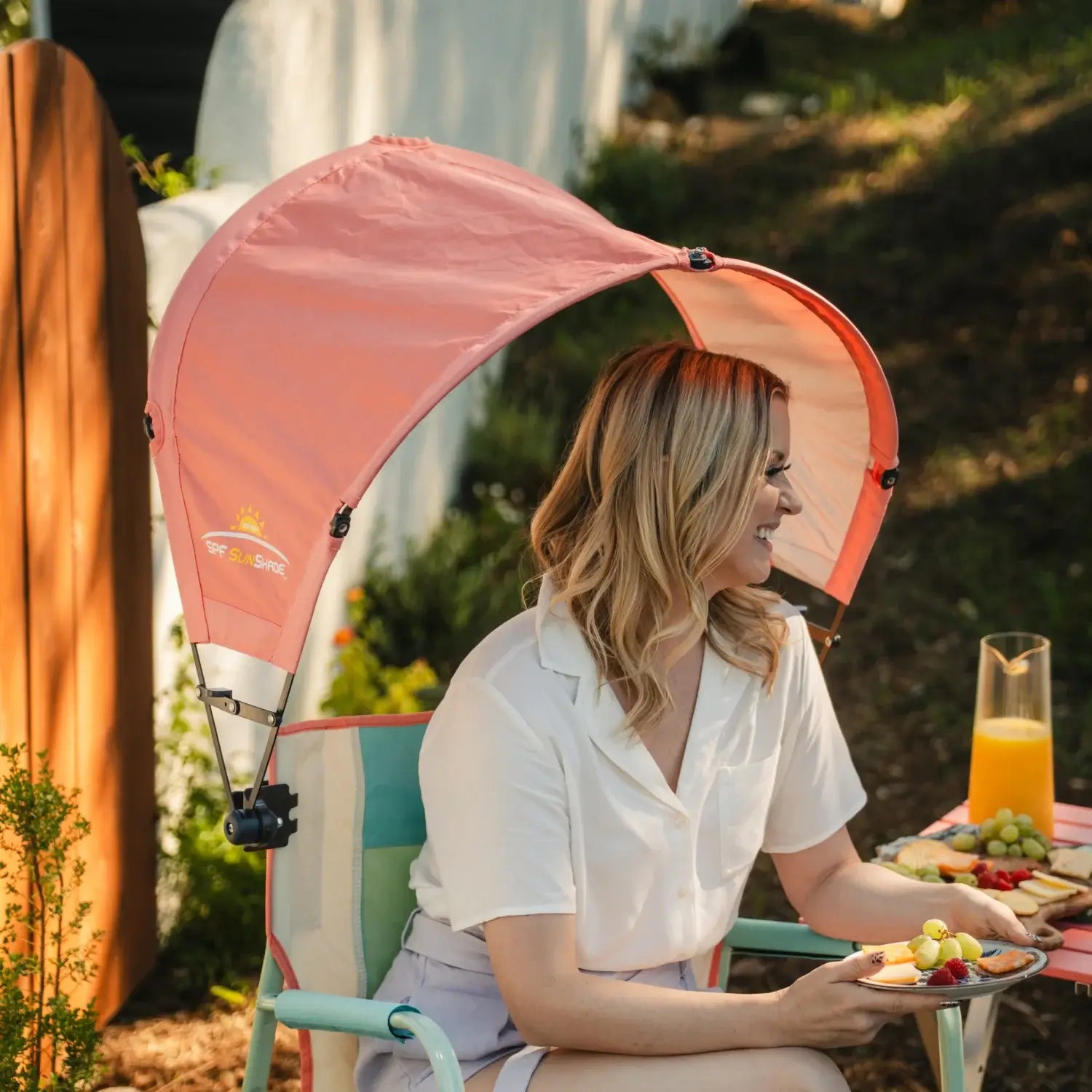 A woman snacking on a charcuterie tray while sitting in an ivory Freestyle Rocker with a coral Sunshade Accessory attached. 