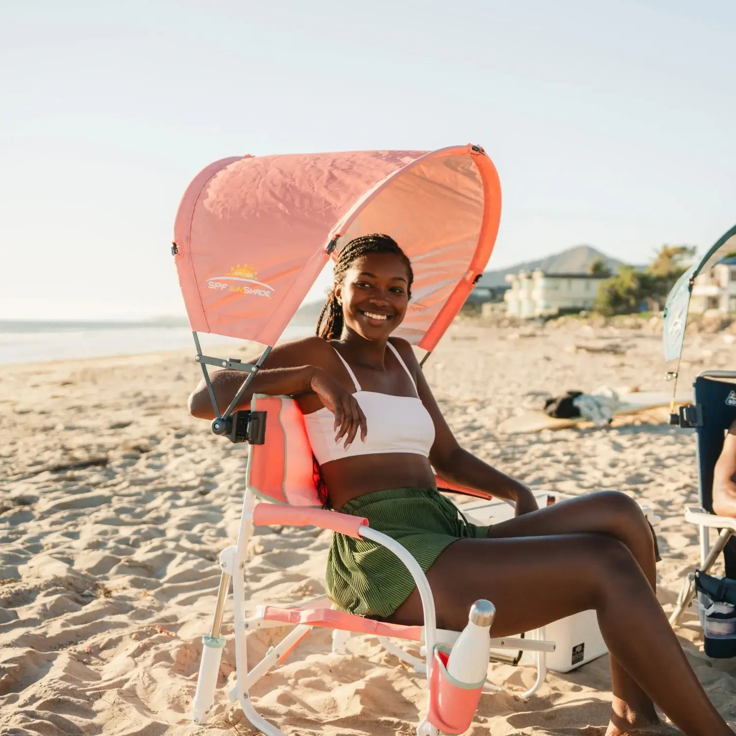 A woman relaxing on a Grab and Go Rocker on a beach with a coral Sunshade Accessory attached. 