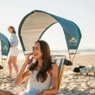 Smiling woman seated under a teal GCI Outdoor SPF SunShade Accessory attached to her beach chair on a sunny shoreline.