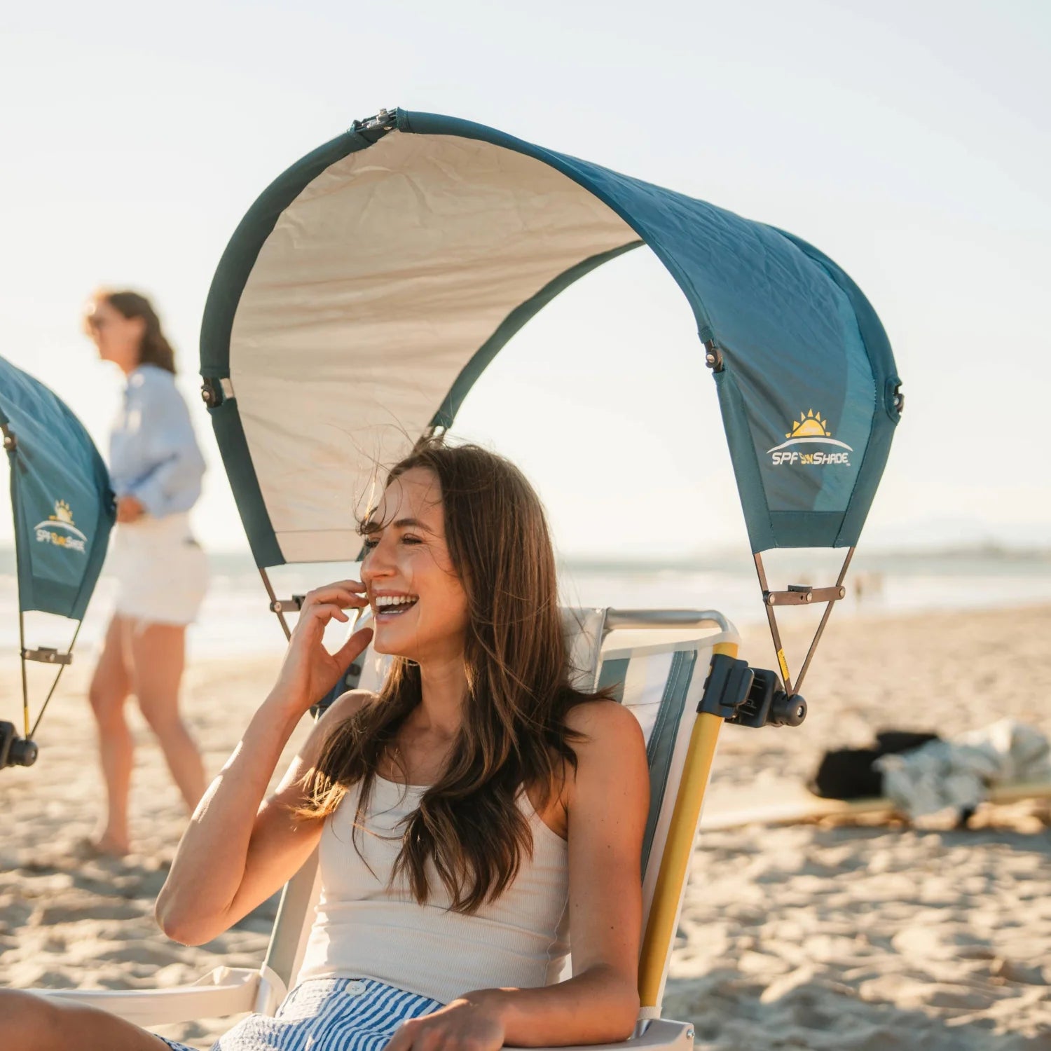 Smiling woman seated under a teal GCI Outdoor SPF SunShade Accessory attached to her beach chair on a sunny shoreline.