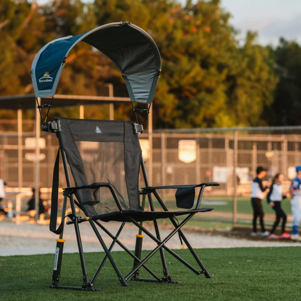 A black GCI outdoor chair with an SPF Sunshade canopy sits on a grassy field at sunset, with kids playing baseball in the background.