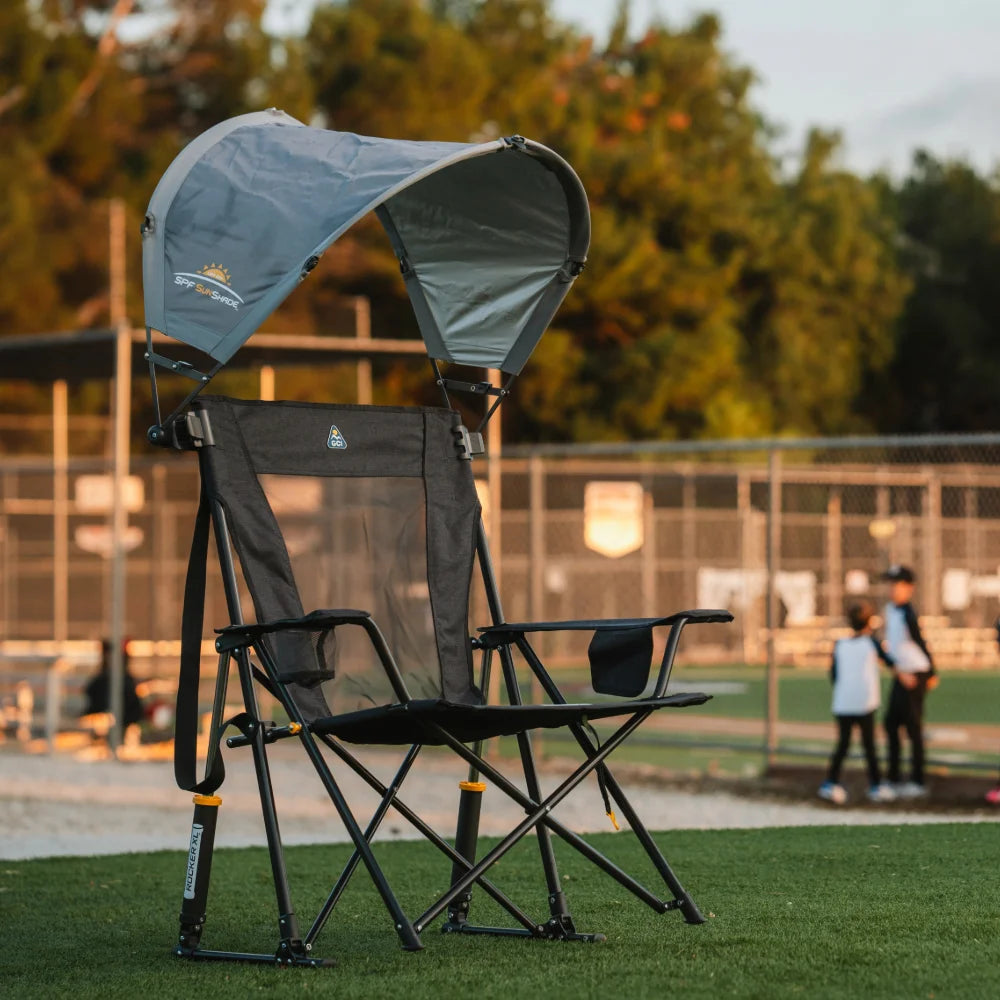 A black GCI outdoor chair with an SPF Sunshade canopy sits on a grassy field at sunset, with kids playing baseball in the background.