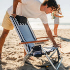 Man setting up the indigo stripe TidalDrift backpack beach chair with wood armrests on the sand.